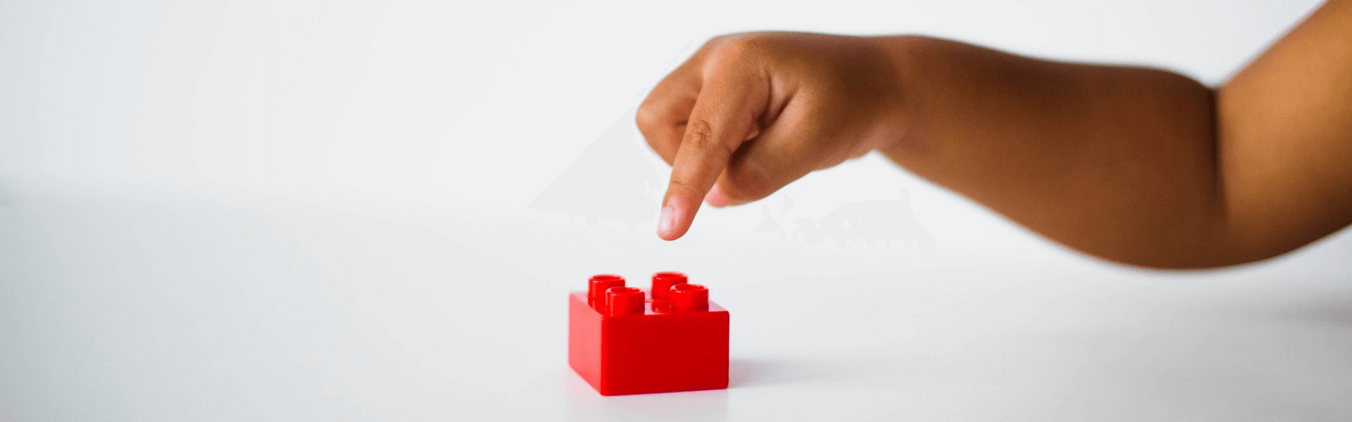 A child extends an index finger over a red plastic building block like he or she is going to touch the top of the building block.