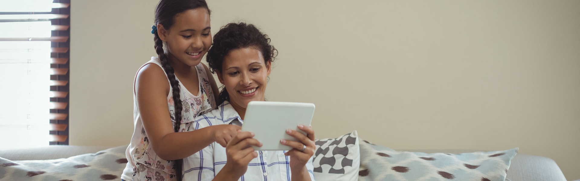 Indoors. On a sofa, a mother and her daughter smile. The mother holds a tablet at chest height. The daughter is kneeling behind the mother's right shoulder and touches the screen of the tablet. 